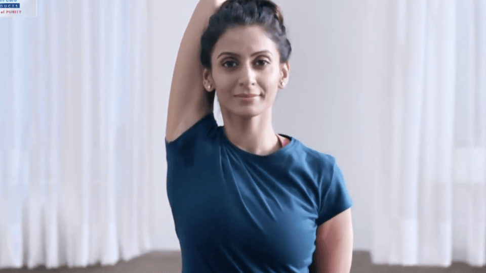 Woman in blue top stretching one arm overhead, calm expression, soft indoor lighting with white curtain background
