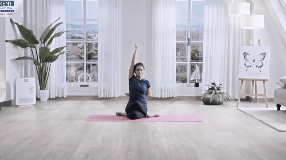 Woman practicing seated yoga pose on a pink mat in a bright living room with large windows and indoor plants