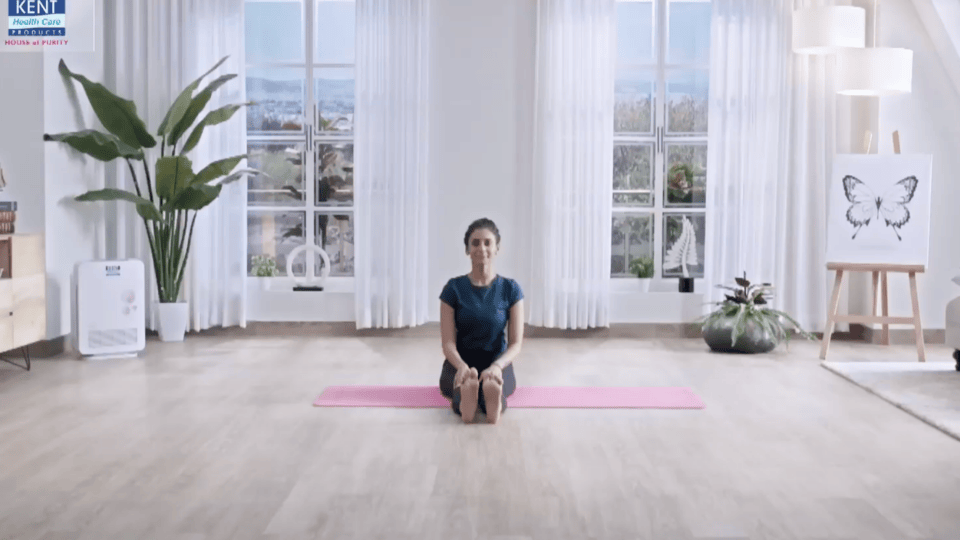 Woman sitting on a yoga mat in a bright living room with large windows, indoor plants, and calm white decor