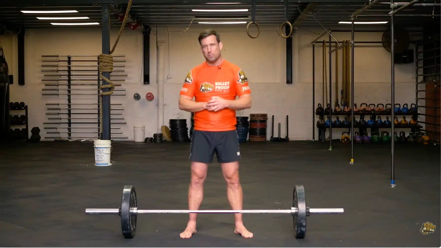 a person stands in a gym next to a barbell with a rack, kettlebells, and rope rings overhead