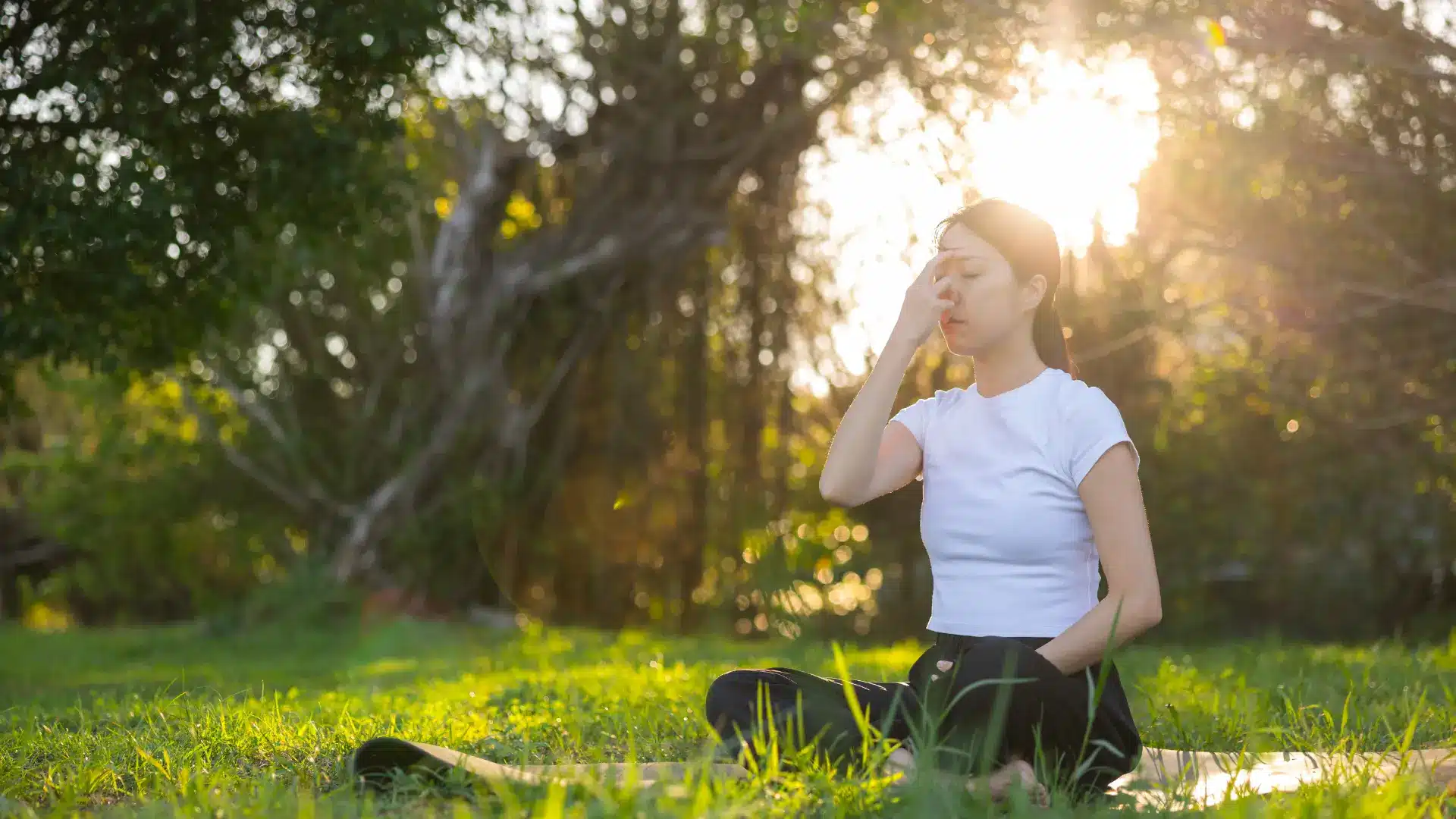 a woman in a white t-shirt sits cross-legged on a mat in a sunlit park, practicing meditative breathing