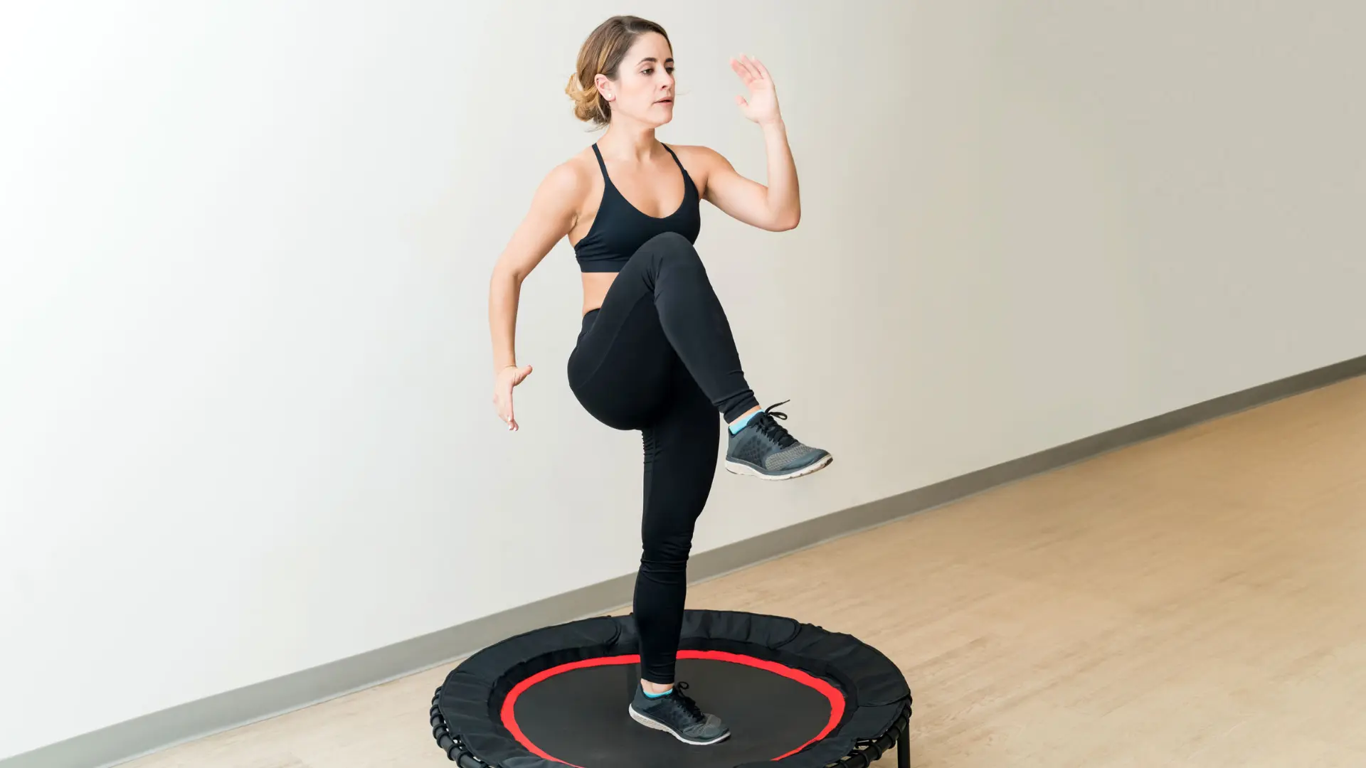 a woman in black athletic wear performs high-knee exercises on a small black and red indoor trampoline