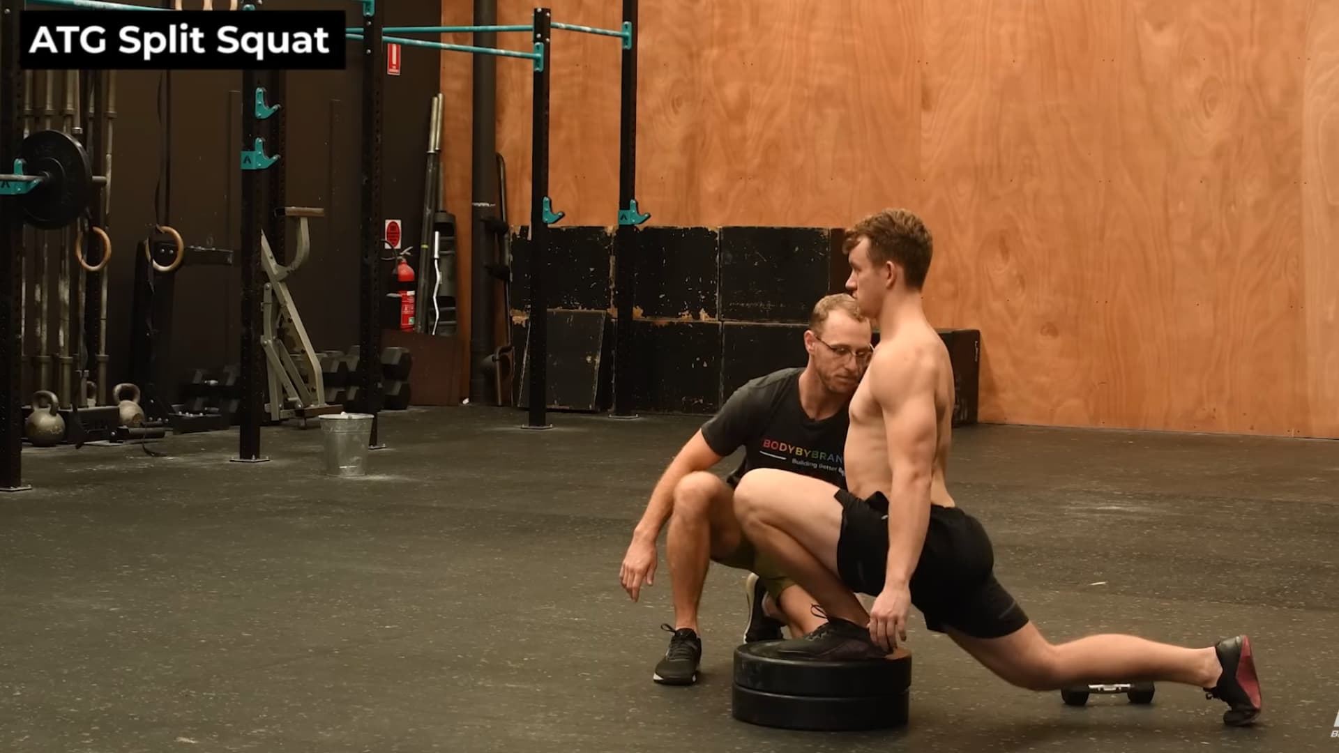 man performs an ATG Split Squat in a gym with his front foot elevated on weight plates. He maintains an upright posture while his coach kneels nearby to observe his form