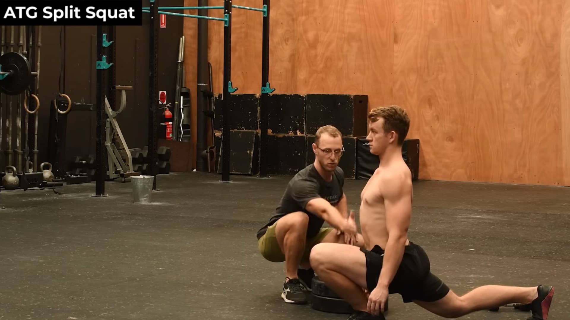 man performs an atg slit squat with his foot elevated on plates while a coach monitors his form