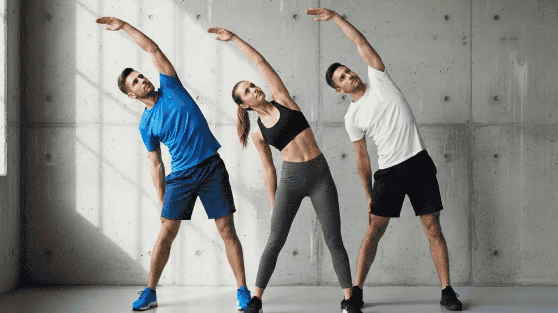 three people in workout gear doing side exercises in a bright gym with a concrete wall.
