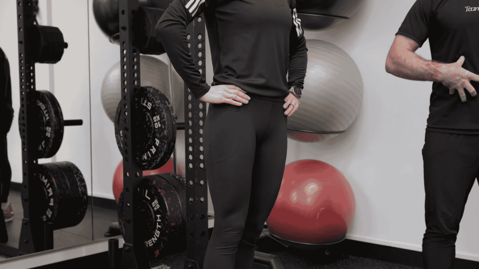 a woman standing on step platform in gym while trainer demonstrates hip positioning for exercise
