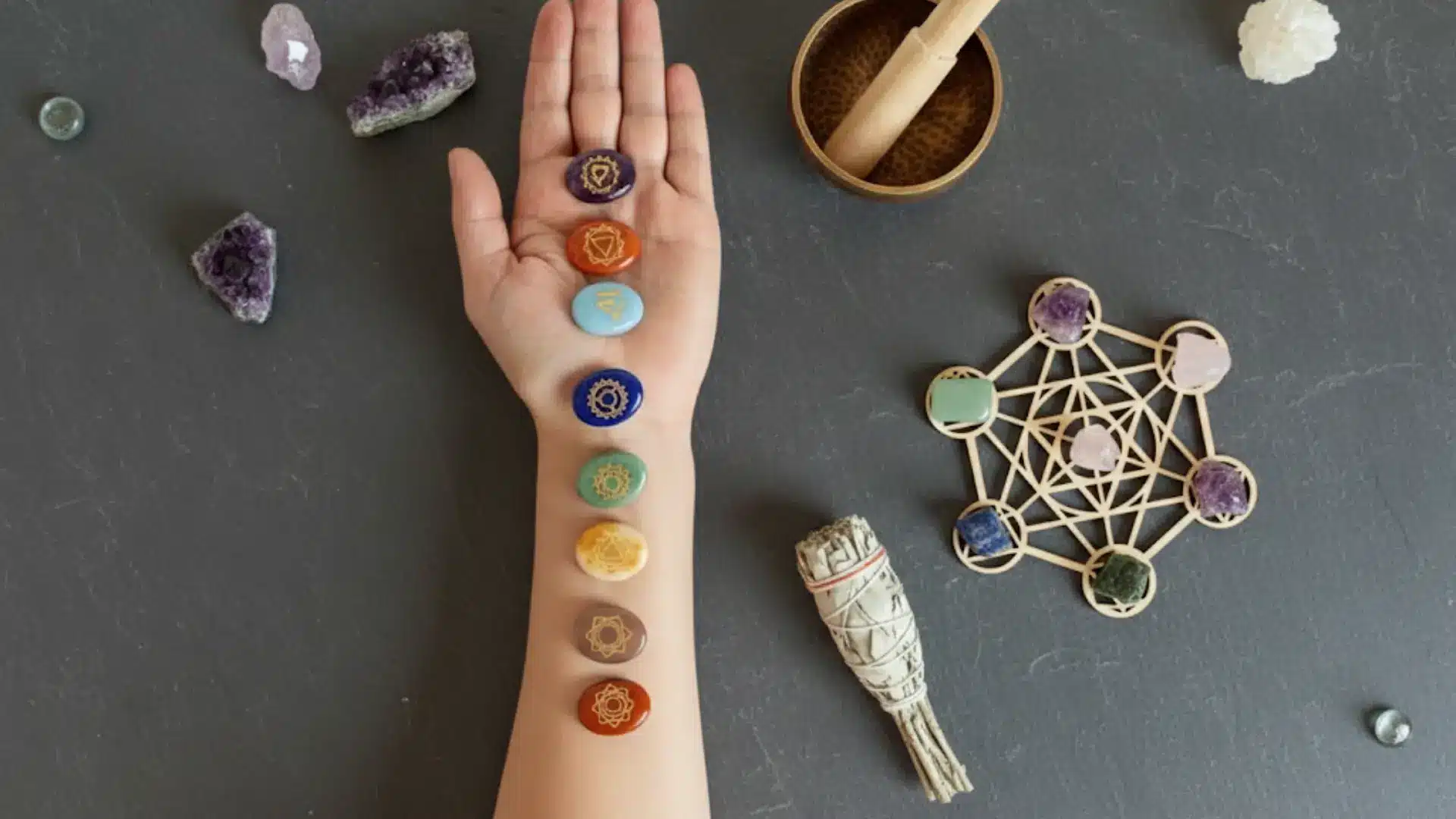 an arm with seven chakra stones, sage, and crystals on a dark grey stone textured background