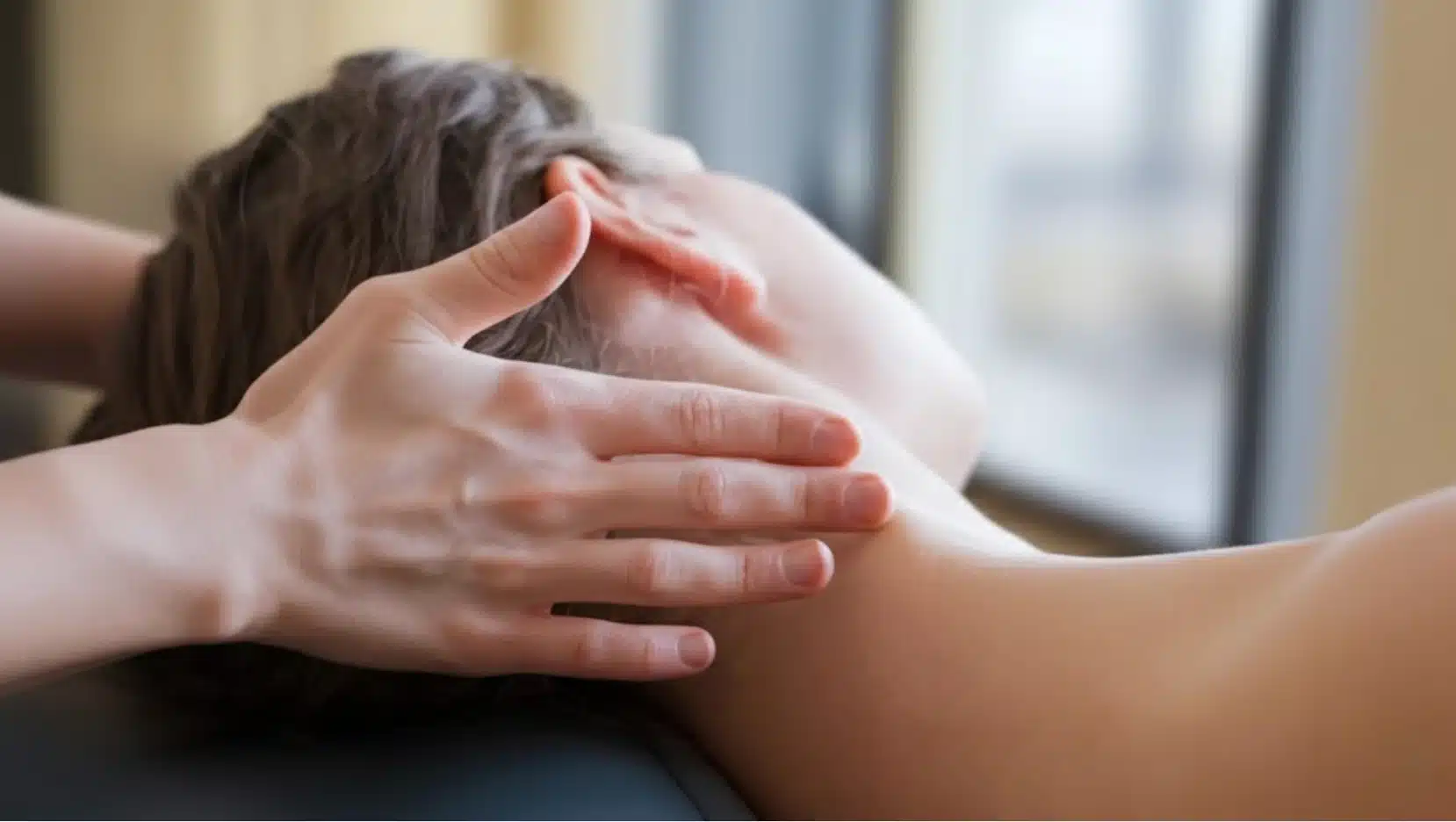 close-up of hands gently massaging a woman's neck as she lies face down on a treatment table near window light