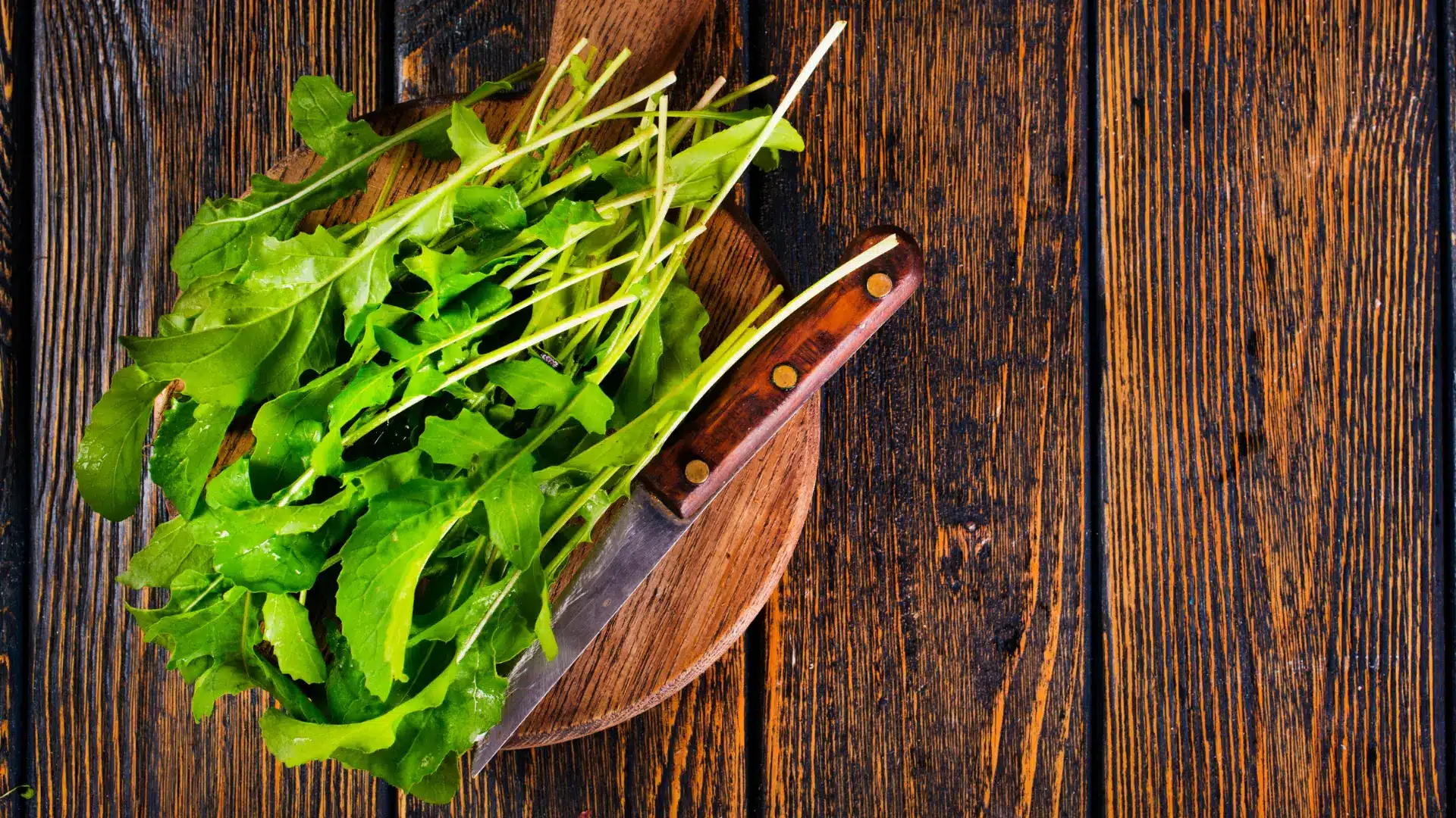 fresh green arugula leaves and a wooden-handled knife resting on a round cutting board over a dark wooden table