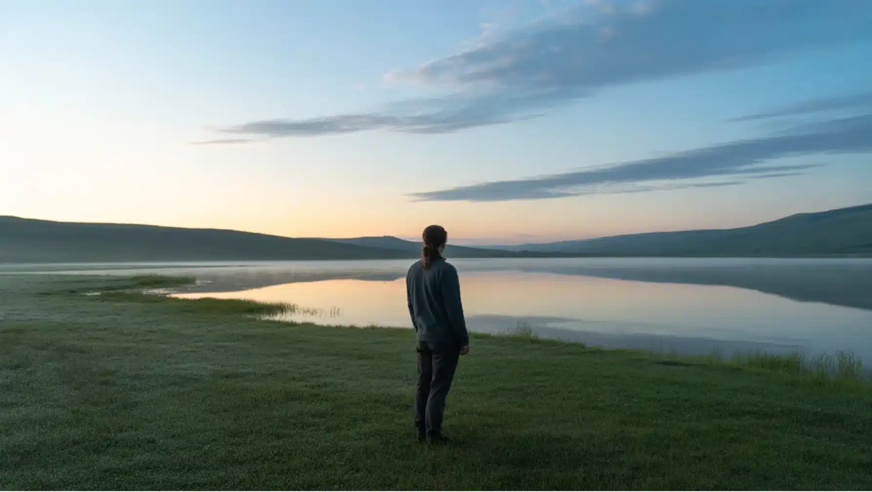 person standing by calm lake at sunrise, gazing across water toward distant hills under soft morning sky light