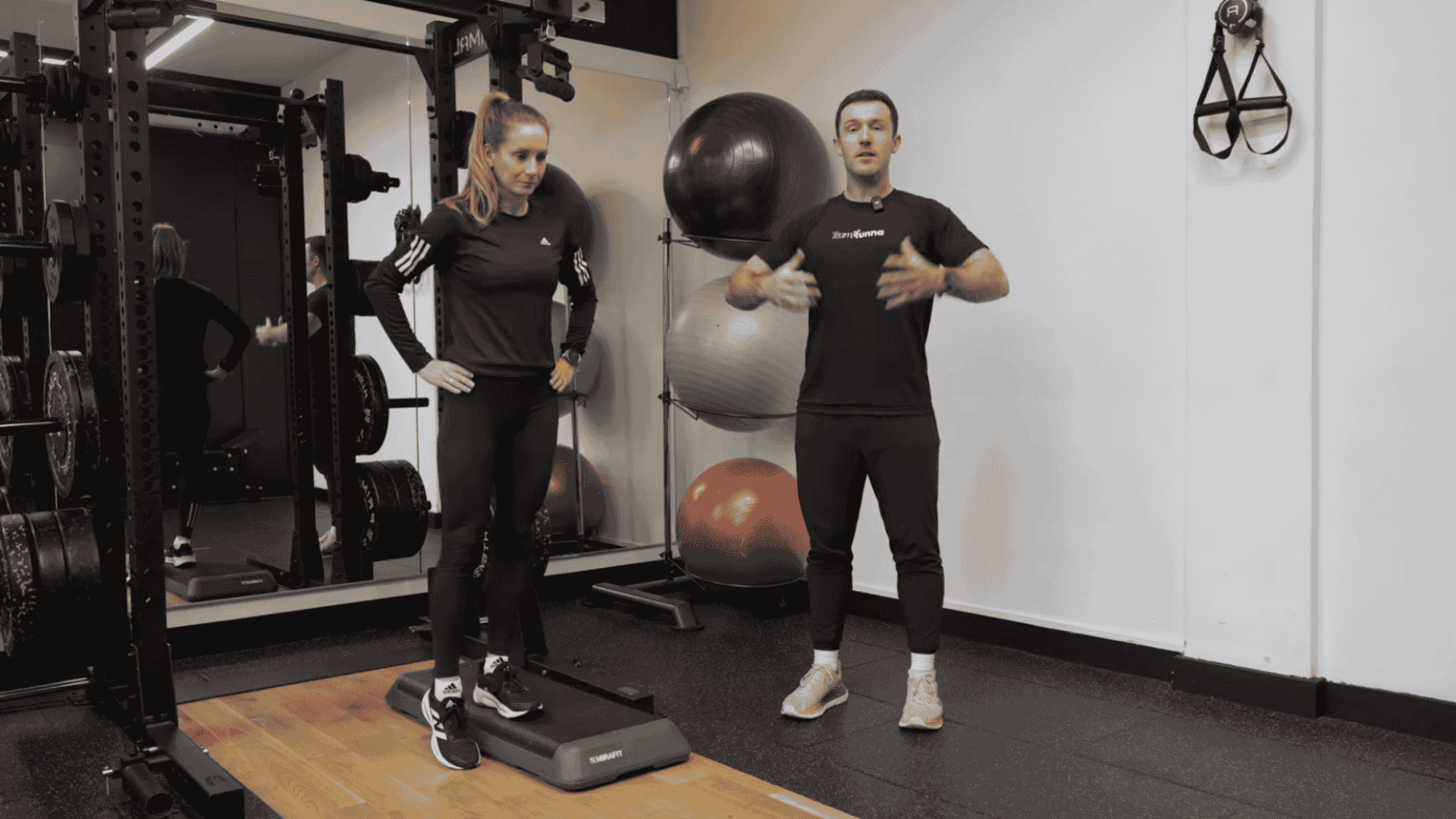 trainer explains hip drop exercise as a woman stands on a step in a gym with fitness equipment behind them