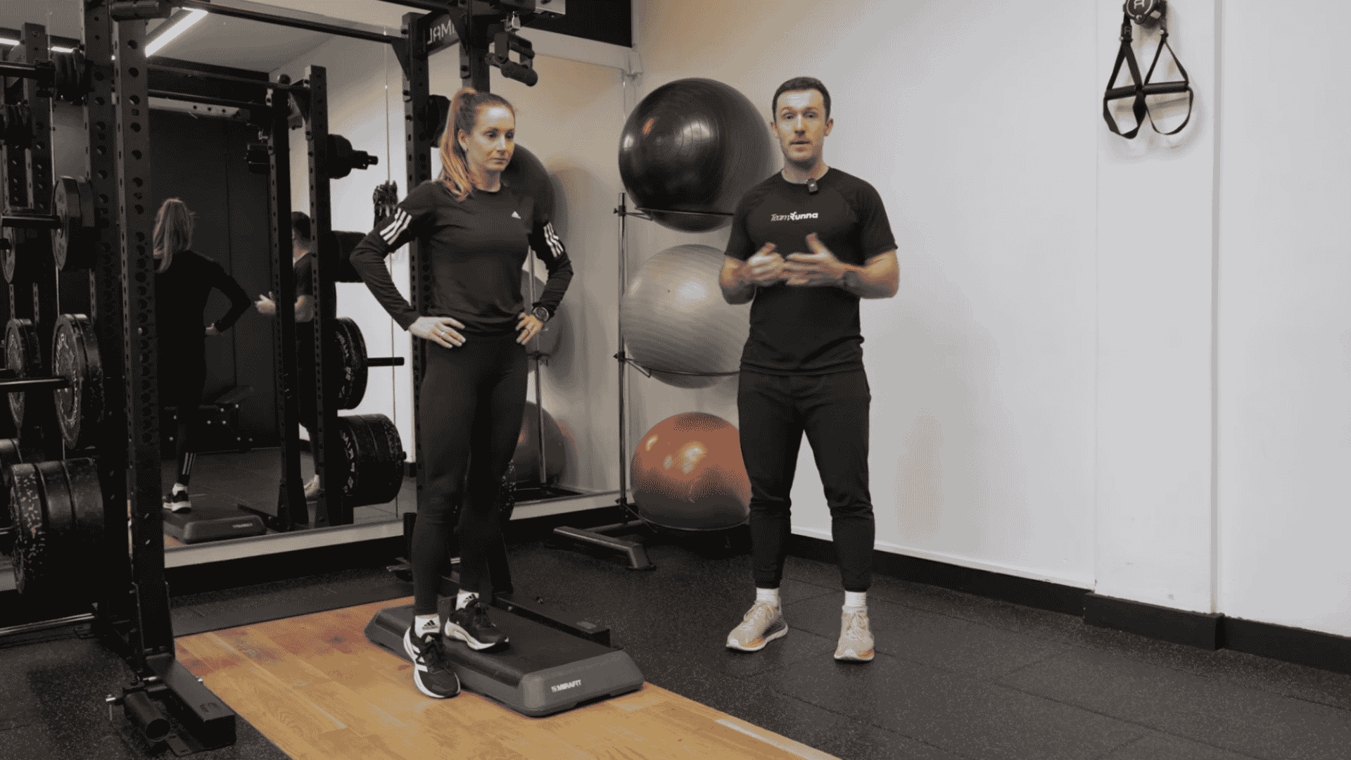trainer instructs woman to drop the hip while standing on step platform in gym with weights and stability balls