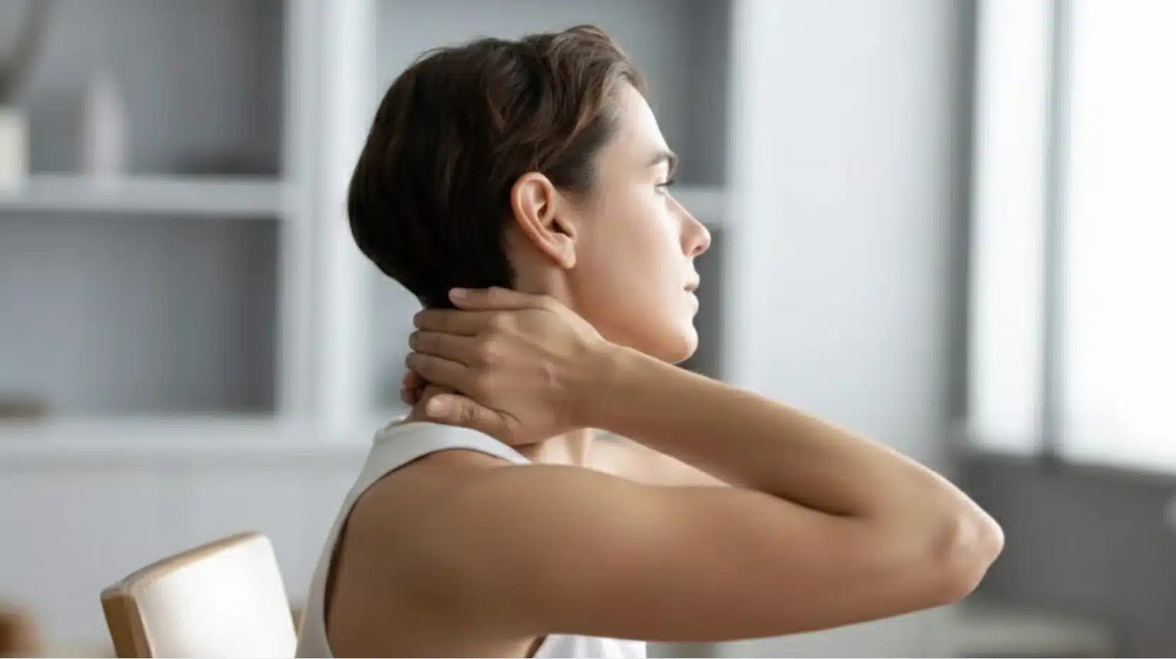 woman sitting indoors holding back of neck with hand, appearing to stretch or relieve tension and discomfort
