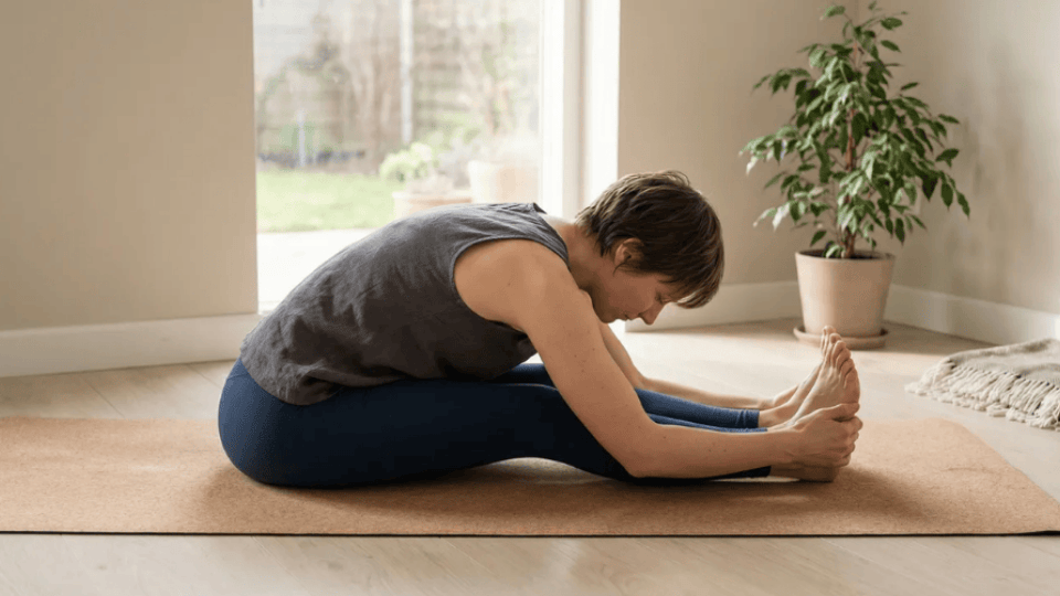 A person practices Seated Forward Fold reaching toward the feet with a calm stretch through the back in a quiet room