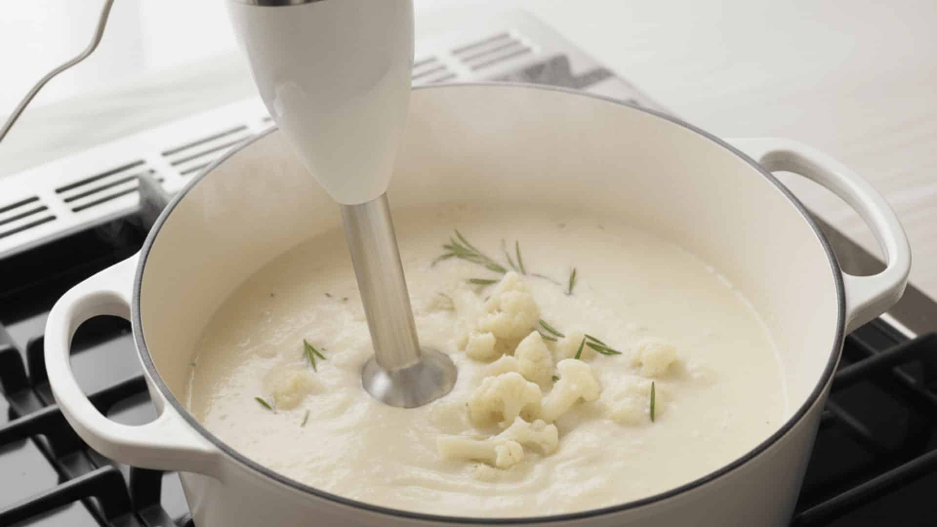 immersion blender blending cauliflower soup in a white dutch oven with rosemary and florets on top