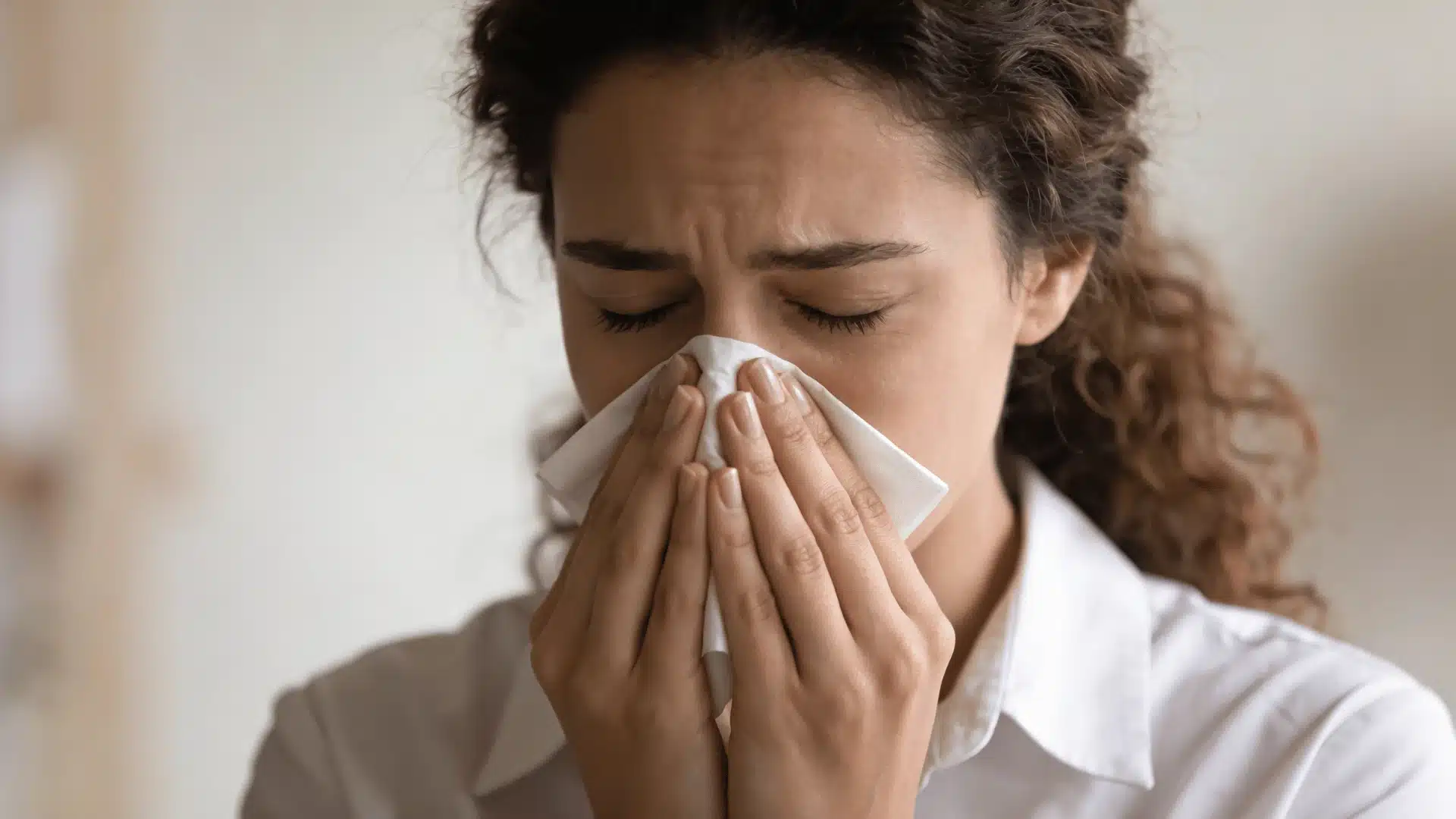 medium shot shows a young woman with curly brown hair, wearing a white collared shirt, blowing her nose with a white tissue