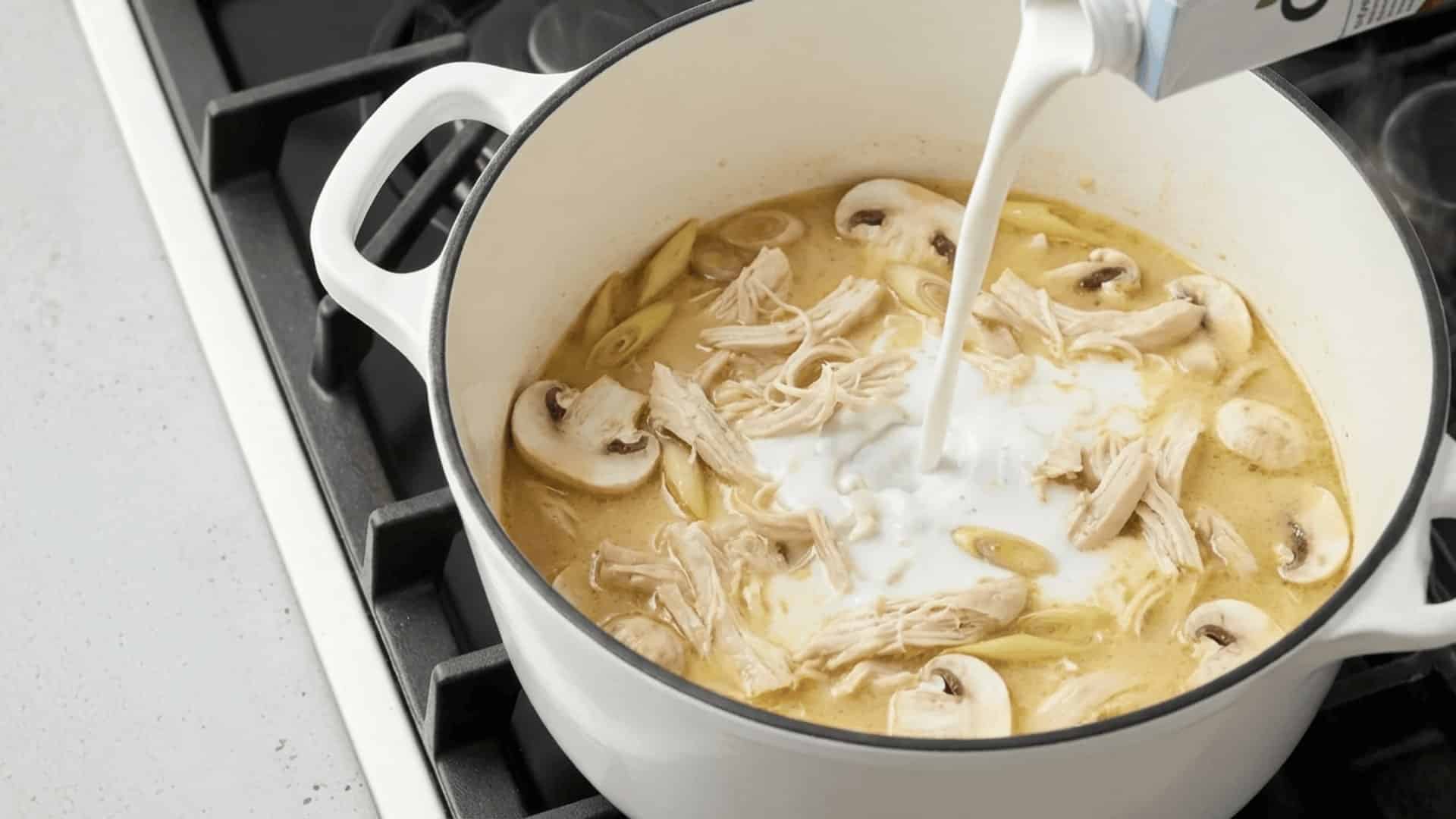 two raw chicken breasts poaching in golden broth with sliced lemongrass and scallions in a white dutch oven
