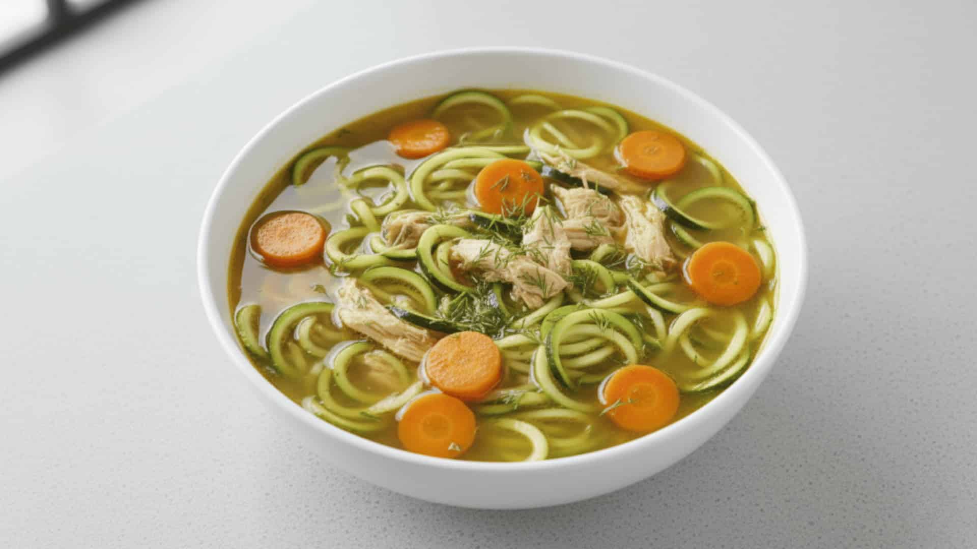 white bowl of shredded chicken soup with spiralized zucchini noodles, sliced carrots, and fresh dill