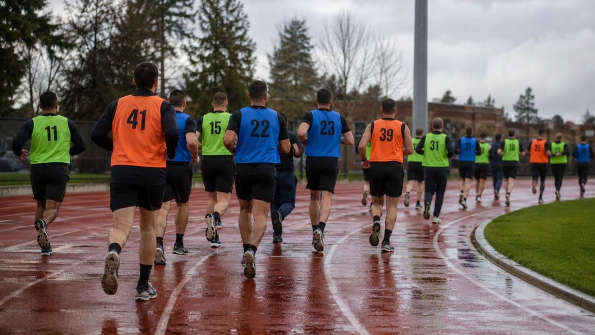 A group of runners wearing brightly colored numbered vests, including orange and blue, sprint on a wet track.