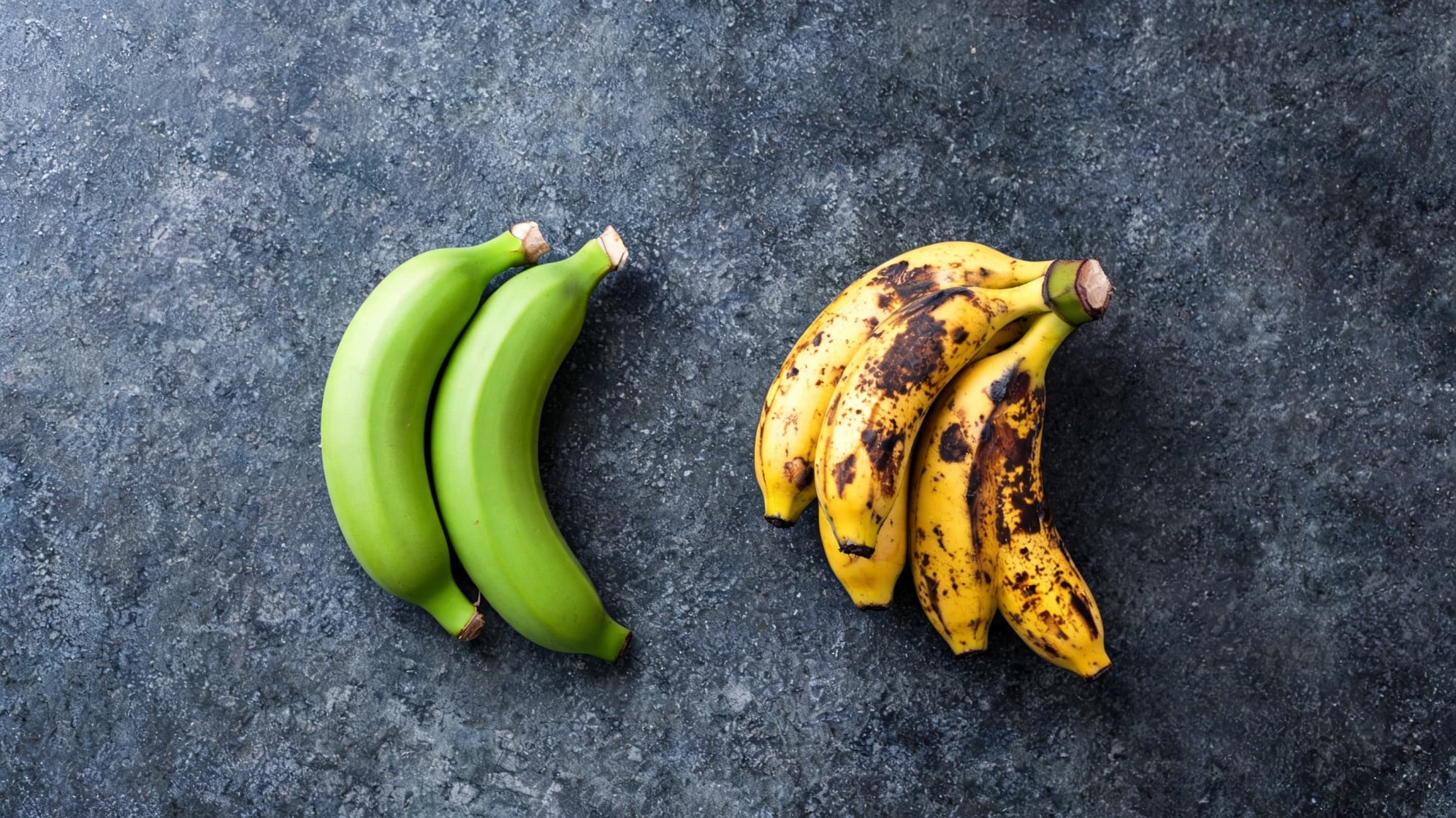 Two clusters of bananas are displayed on a dark. On the left, unripe green bananas and on the right, four ripe