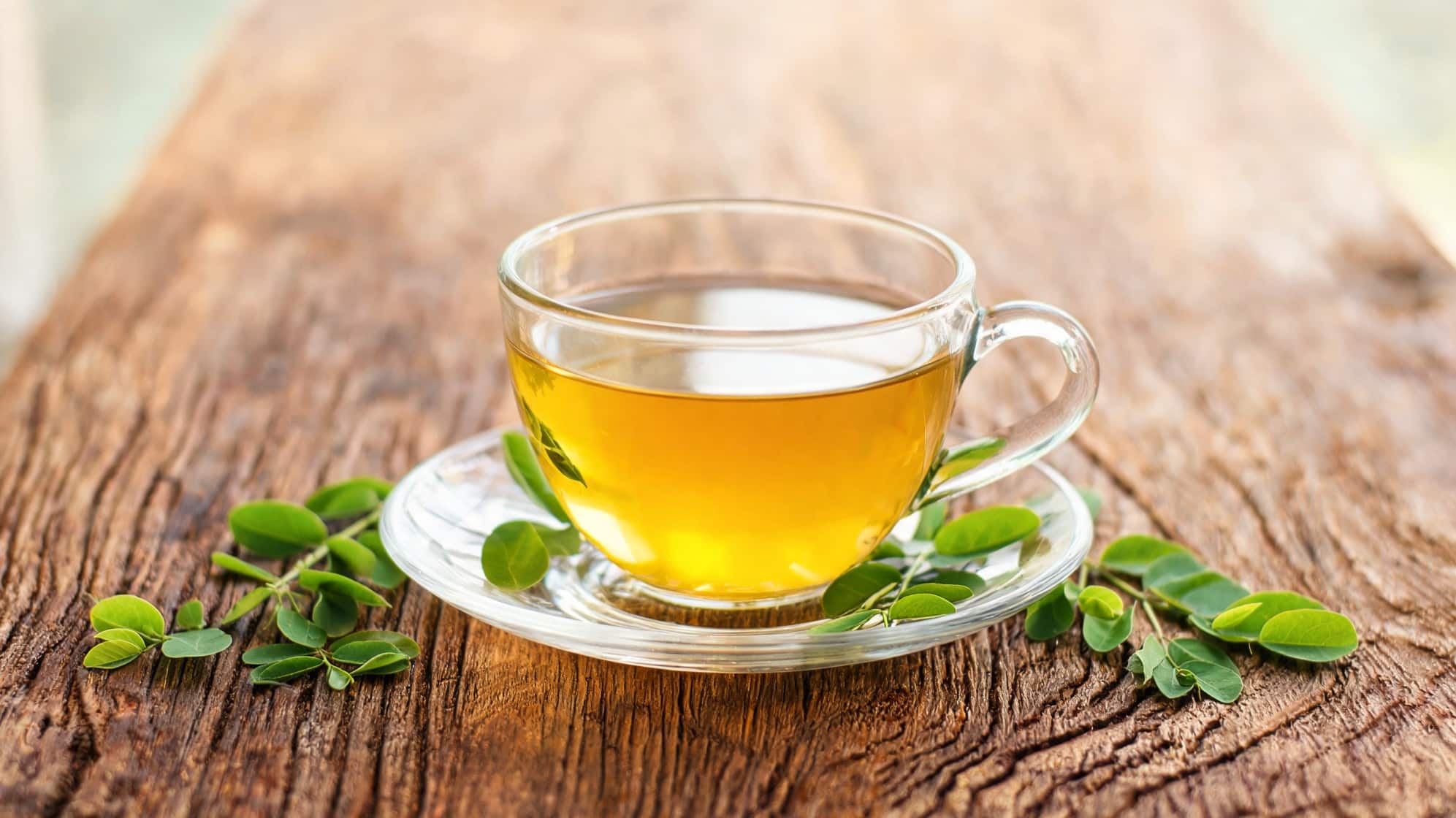 A glass cup of moringa tea sits on a rustic wooden table, surrounded by fresh moringa leaves