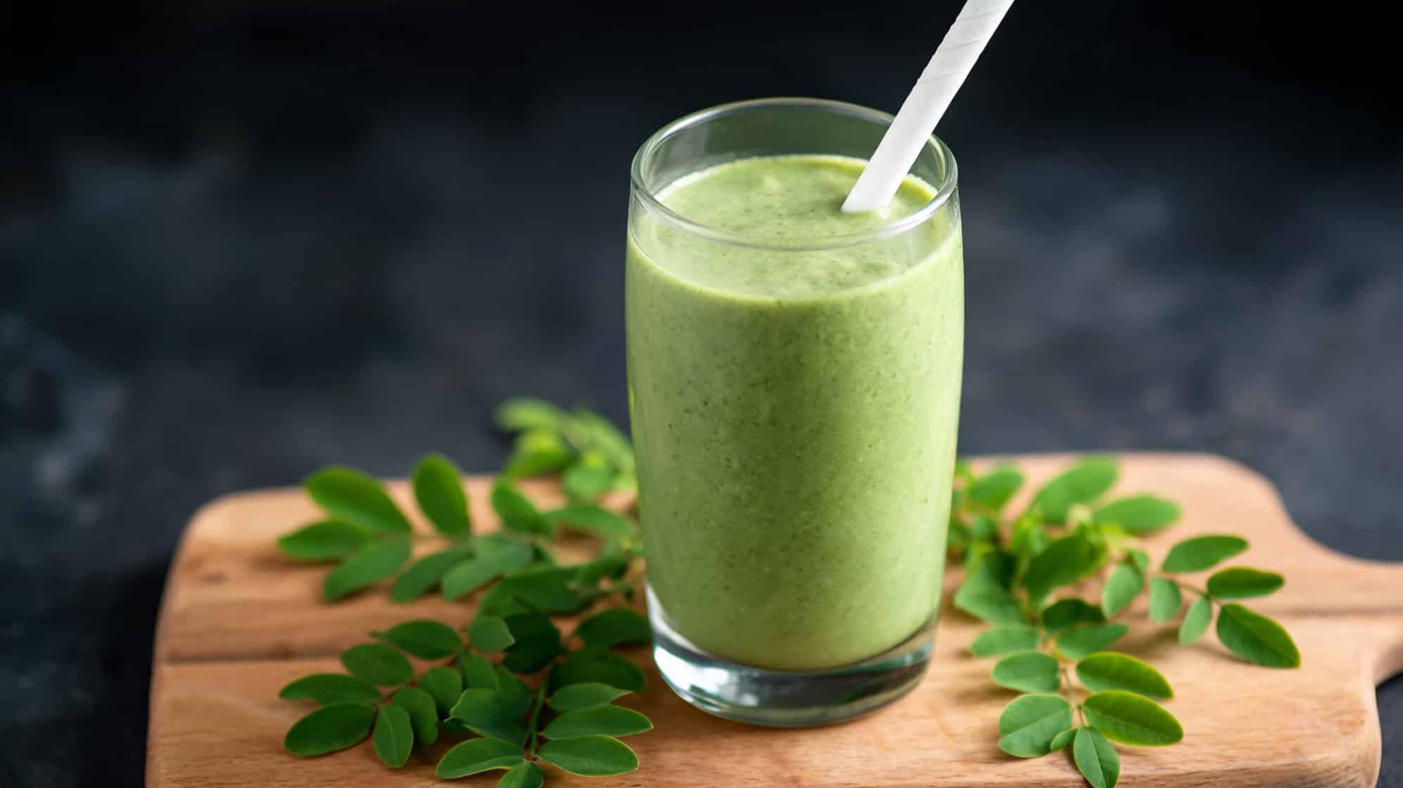 A moringa smoothie in a glass with a straw sits on a wooden board, surrounded by fresh moringa leaves