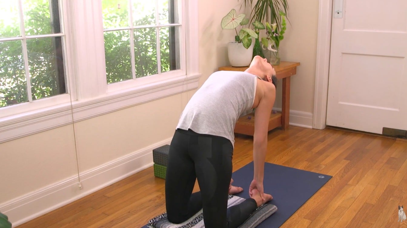 A person in a white tank top and black leggings performs a yoga camel pose on a mat in a sunlit room