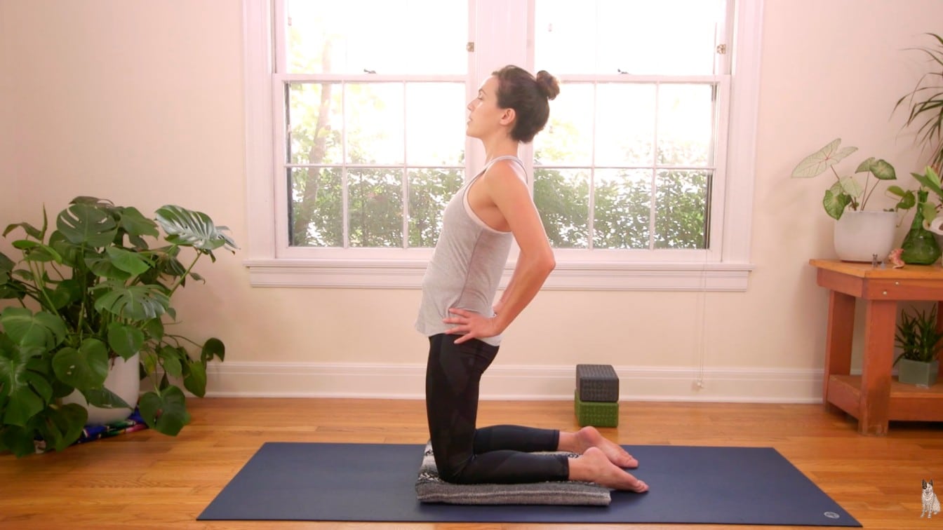 A person in athletic wear practices yoga, sitting on their heels with hands on hips, in a bright room