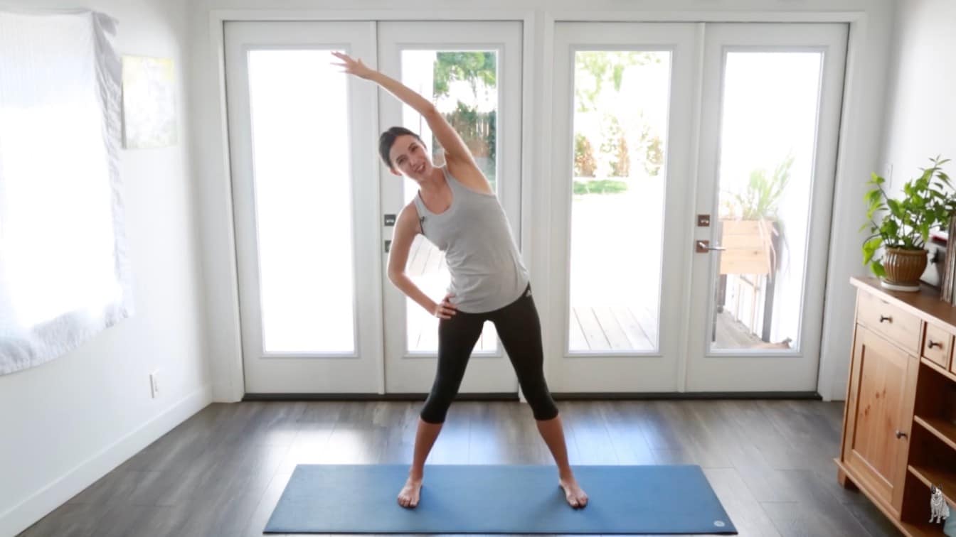 a woman in workout attire performs a side stretch on a yoga mat in a bright room with large glass doors