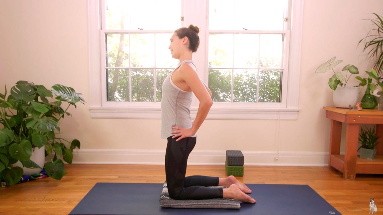 A woman performs yoga in a serene room, kneeling on a mat with hands on hips, set against a backdrop of a window