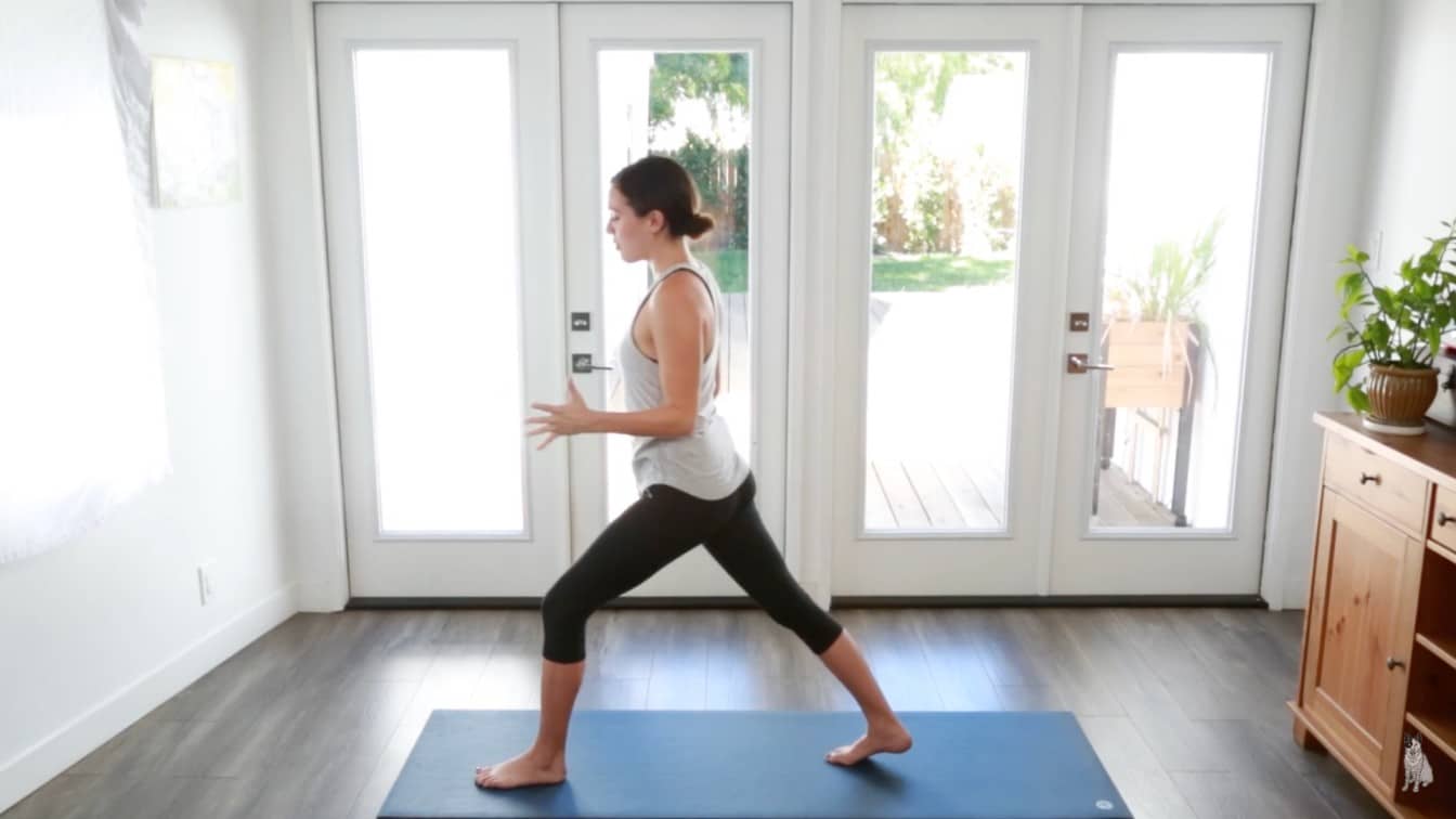 a woman practices yoga in a bright room with wooden floors standing in a lunge pose on a blue mat