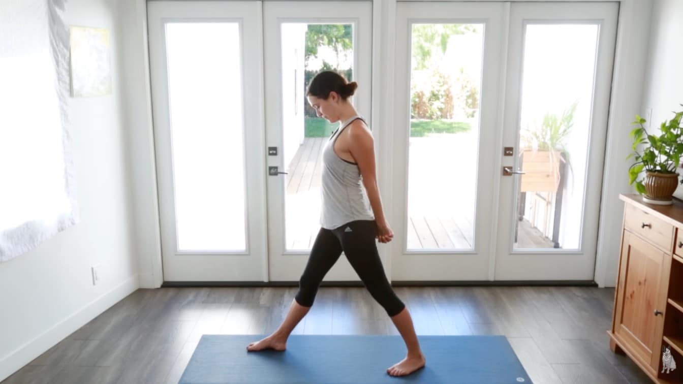 a woman practicing yoga in a bright room, standing in a forward bend pose on a blue mat
