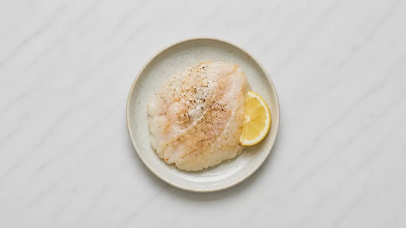 cooked tilapia fillet on a white plate with lemon on marble