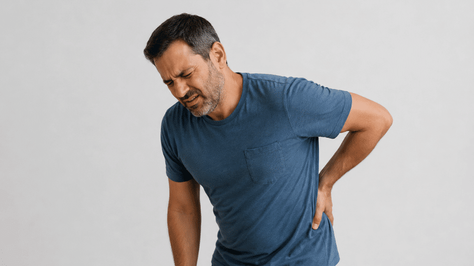 middle aged man holding lower back in pain against a plain editorial style background