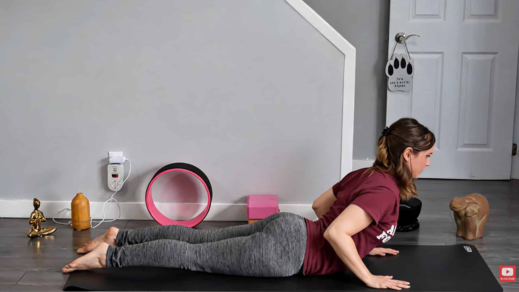 woman performing baby cobra pose on a black yoga mat with a pink yoga wheel and block in the background