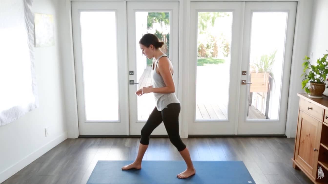 woman practices yoga in a bright room, standing on a blue mat. She wears a white tank top and black leggings