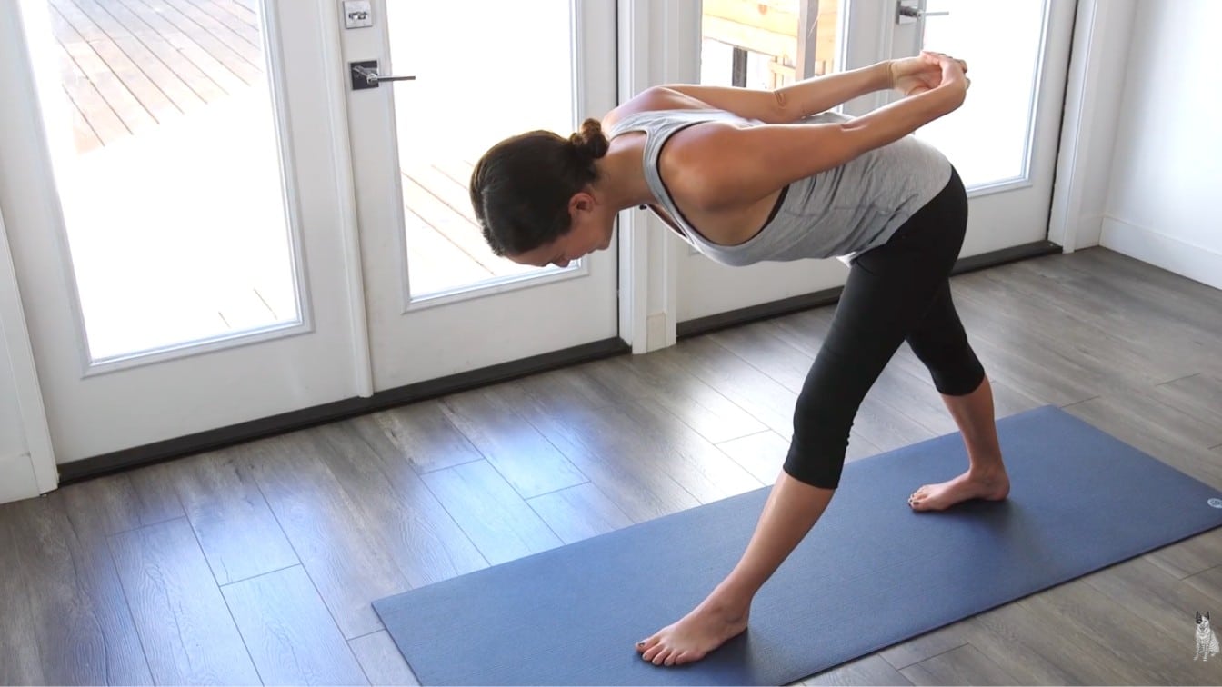 woman practices yoga on a mat in a sunlit room. She's in a forward bend, legs apart, and hands clasped behind her back