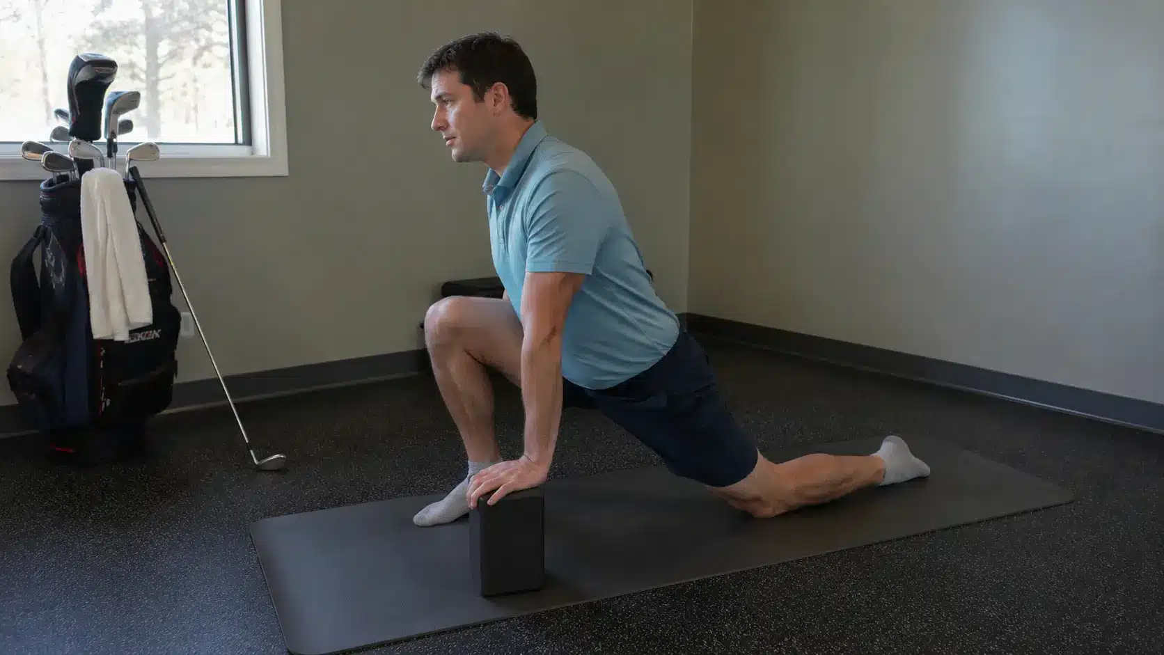 a regular golfer is using a yoga block during a supported low lunge in a simple fitness corner with a golf bag towel and club nearby to show safer stretching without forcing the pose