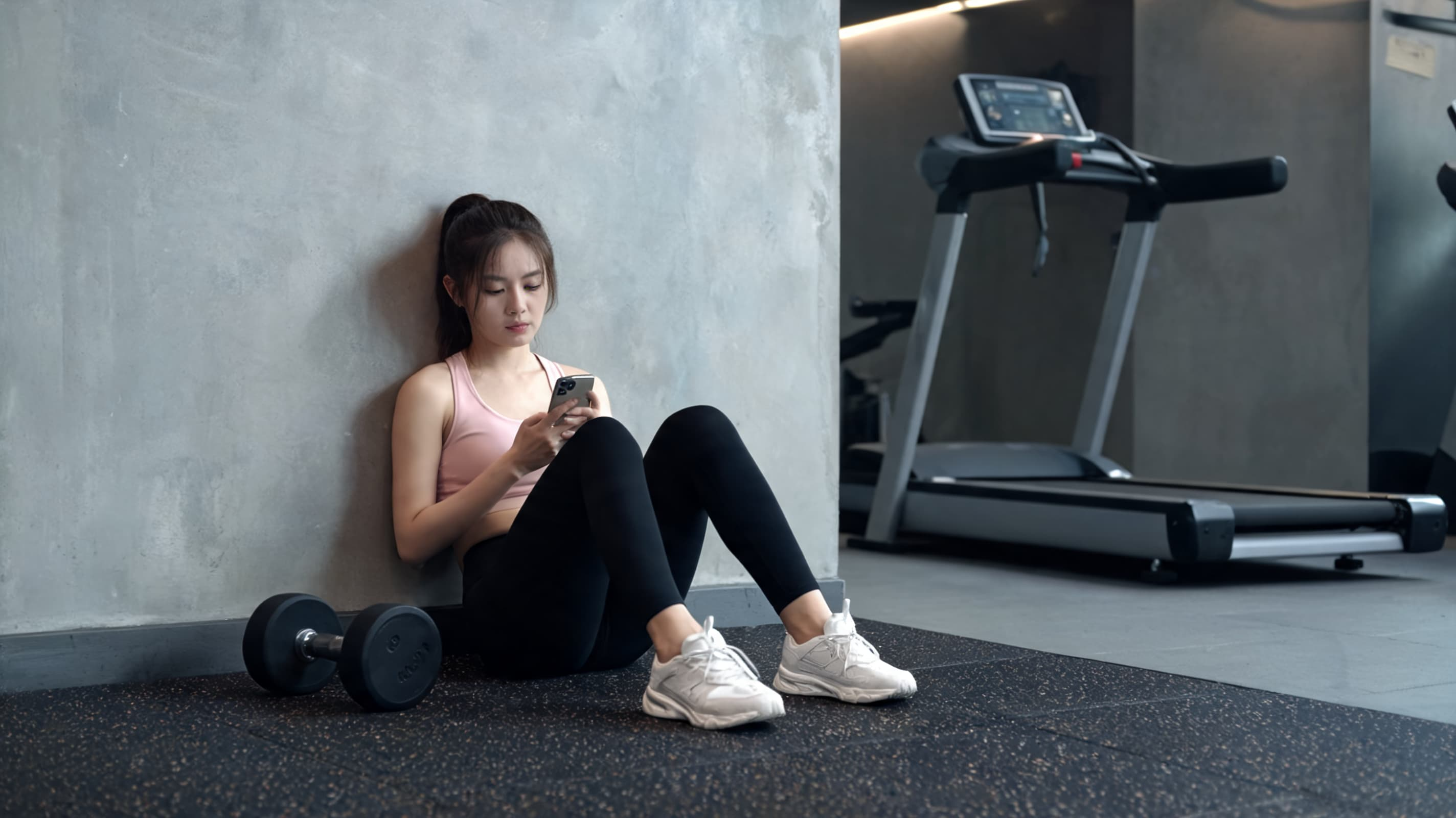 Woman in workout clothes sitting against a gym wall looking at her phone