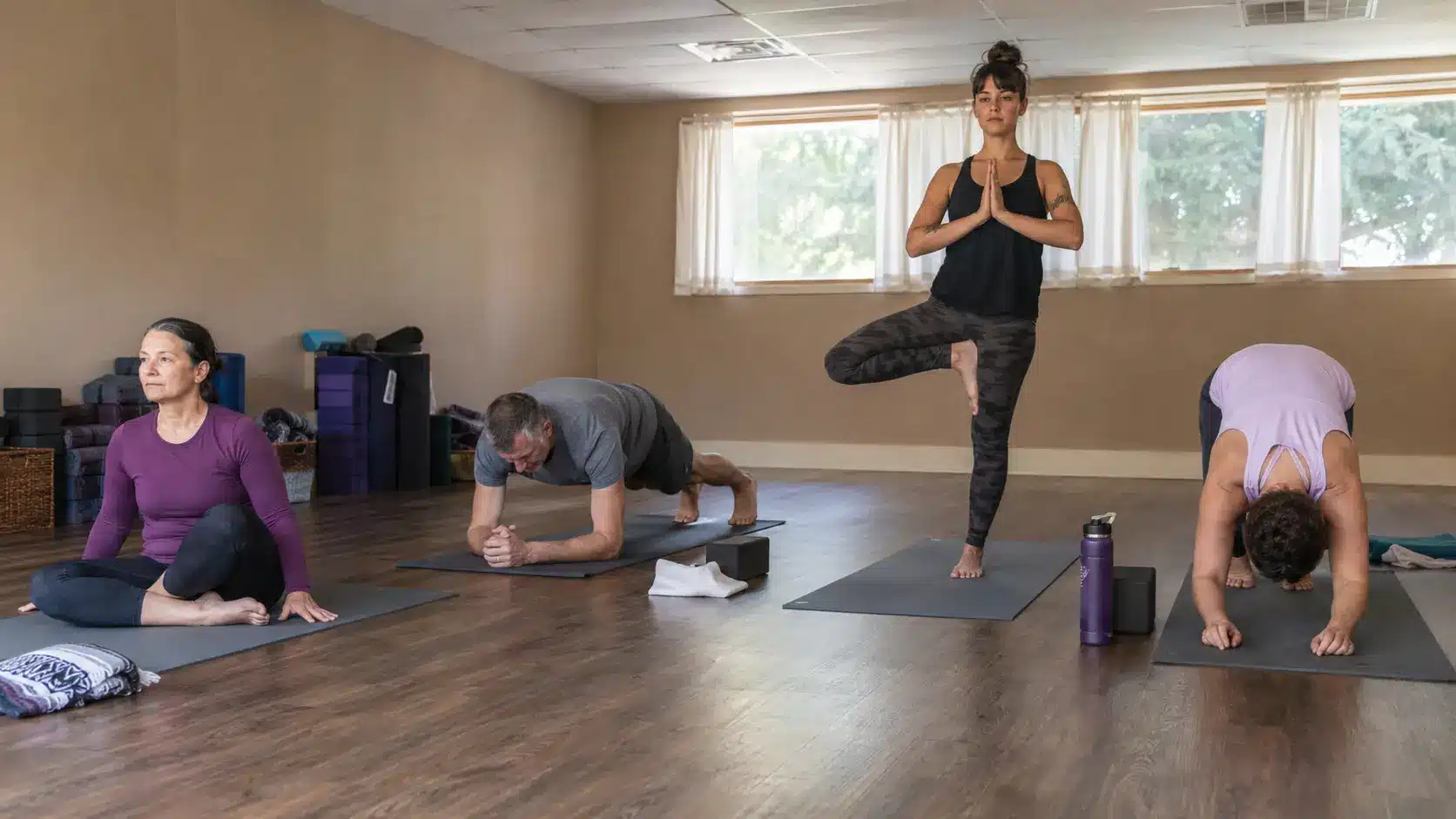 four people practice different stages of a short group yoga routine in a simple studio with mats towels and water bottles nearby