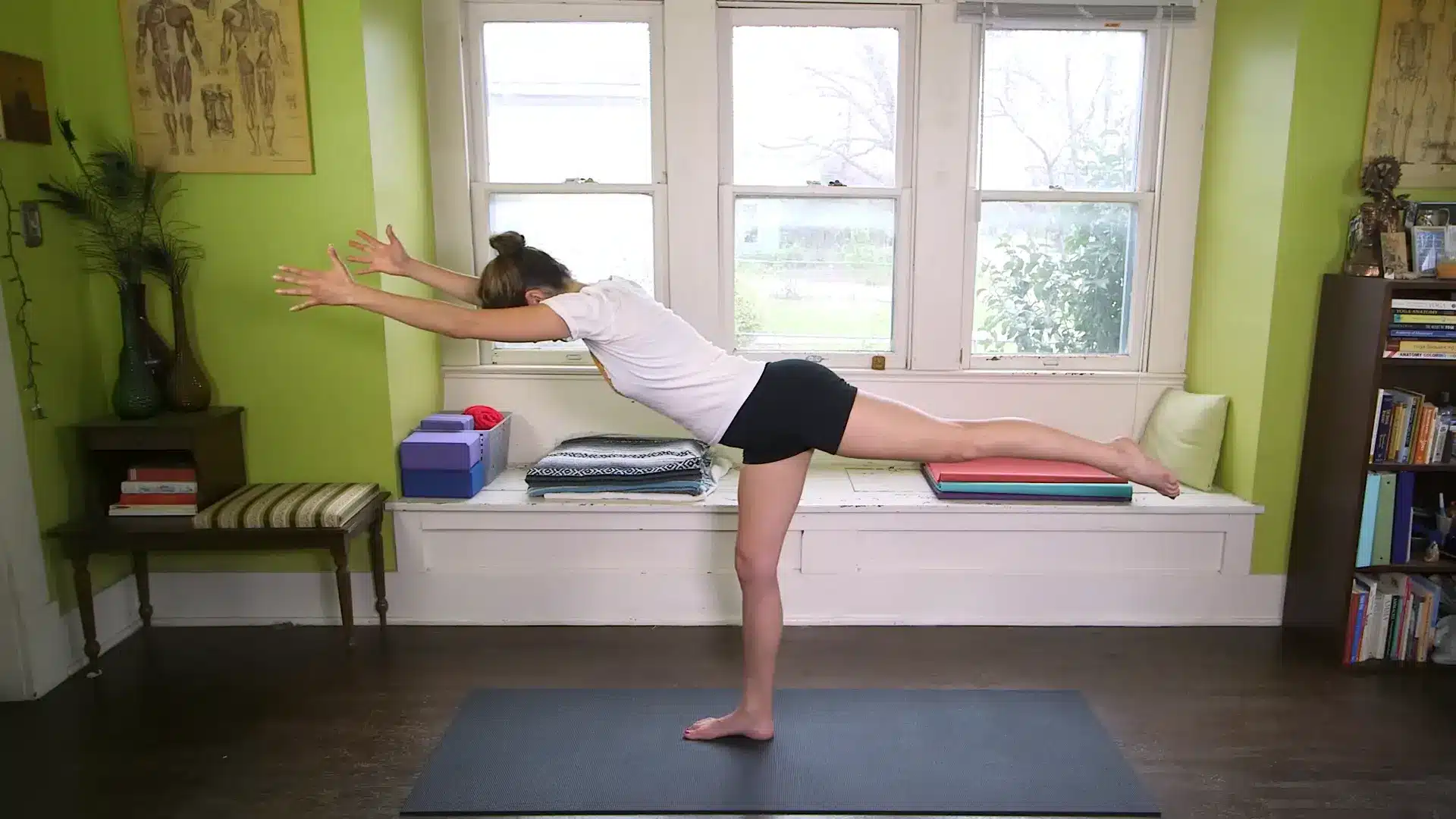 person holding full warrior three pose with arms forward and back leg lifted in a bright home yoga room