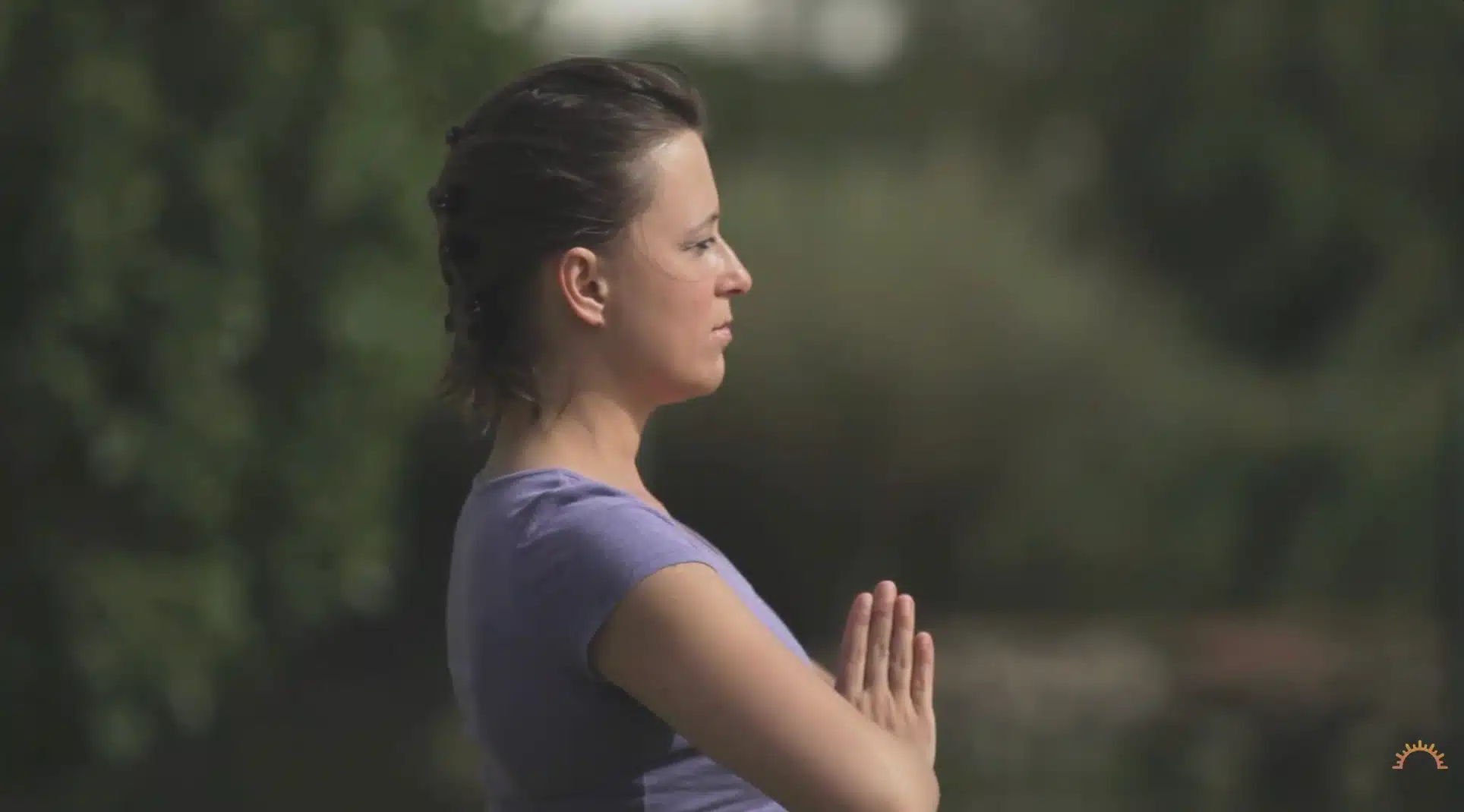 woman in side profile holds prayer pose outdoors with calm focus against a blurred background