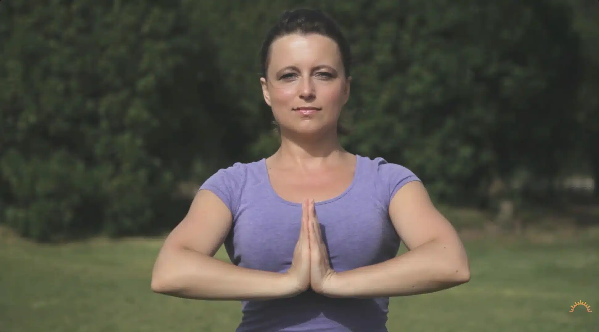 woman holds prayer pose outdoors with palms together in a calm green setting