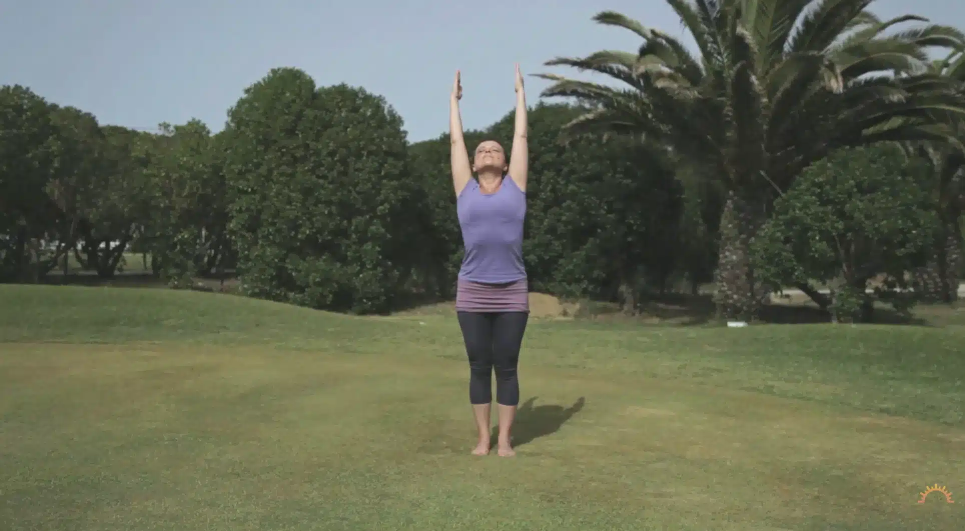 woman stretches both arms upward outdoors during a calm sun salutation practice on grass