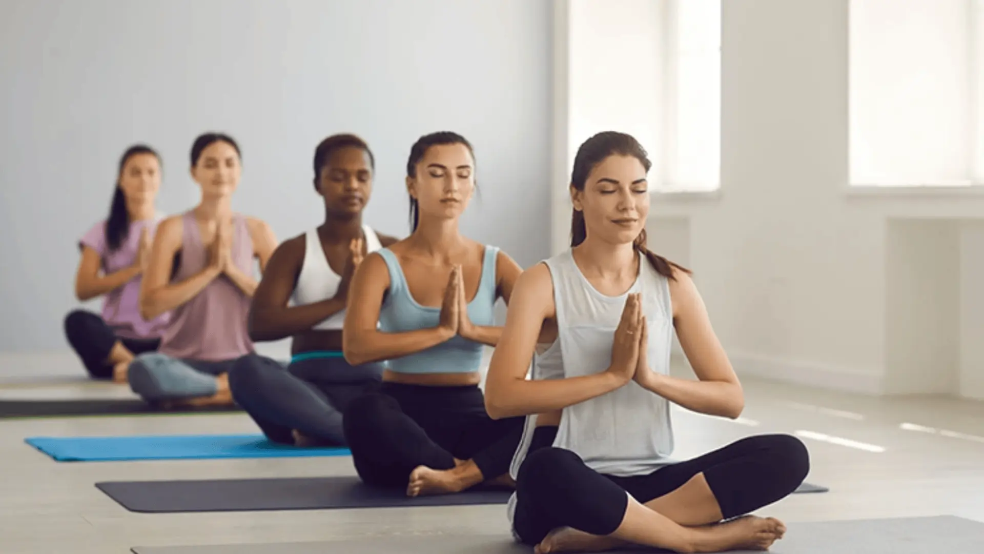 diverse group of women sit cross-legged in a bright studio, meditating with their eyes closed and hands in prayer