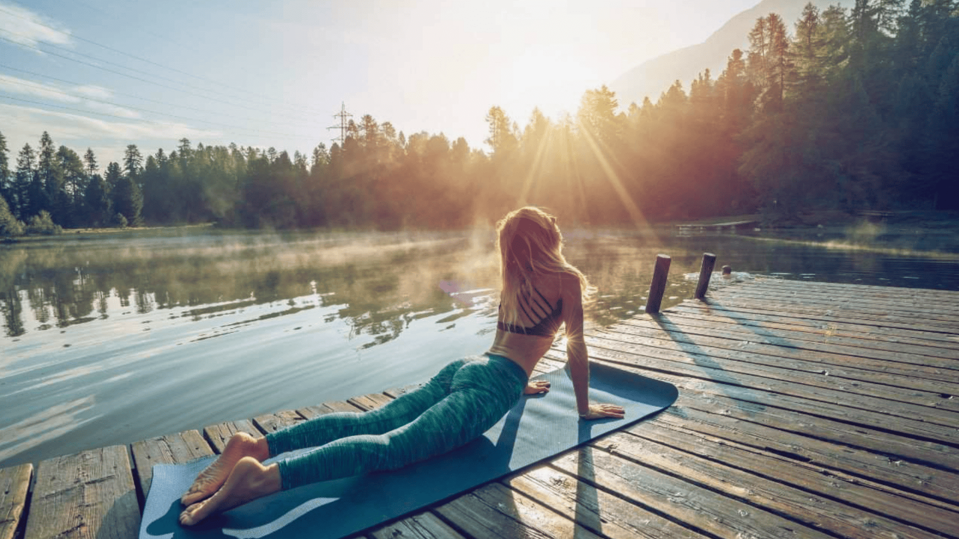 woman practices an upward-facing dog pose on a wooden dock by a misty, sunlit lake during sunrise