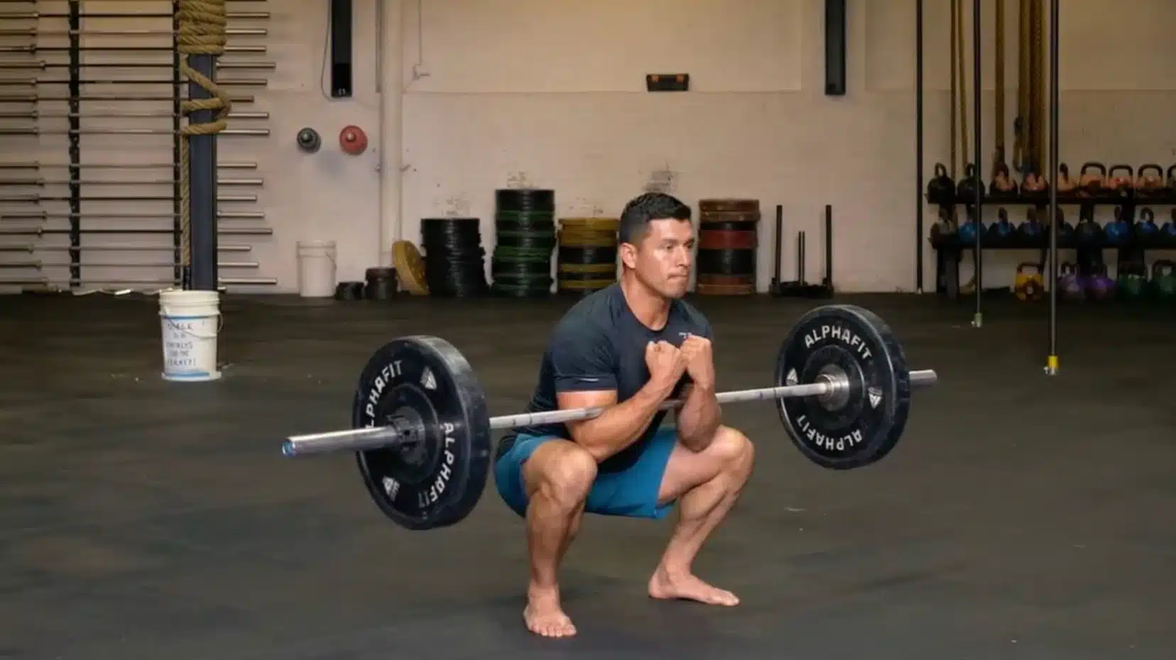 a man performs a zercher squat with a barbell racked at the elbows, lifting in a gym with boxing bags in the background