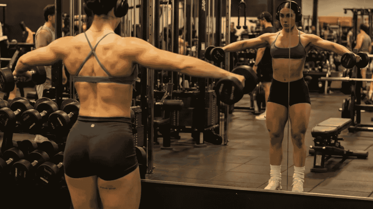 a person performing a dumbbell lateral+raise+in+a+gym,+focusing on+their shoulders, with gym equipment in the background