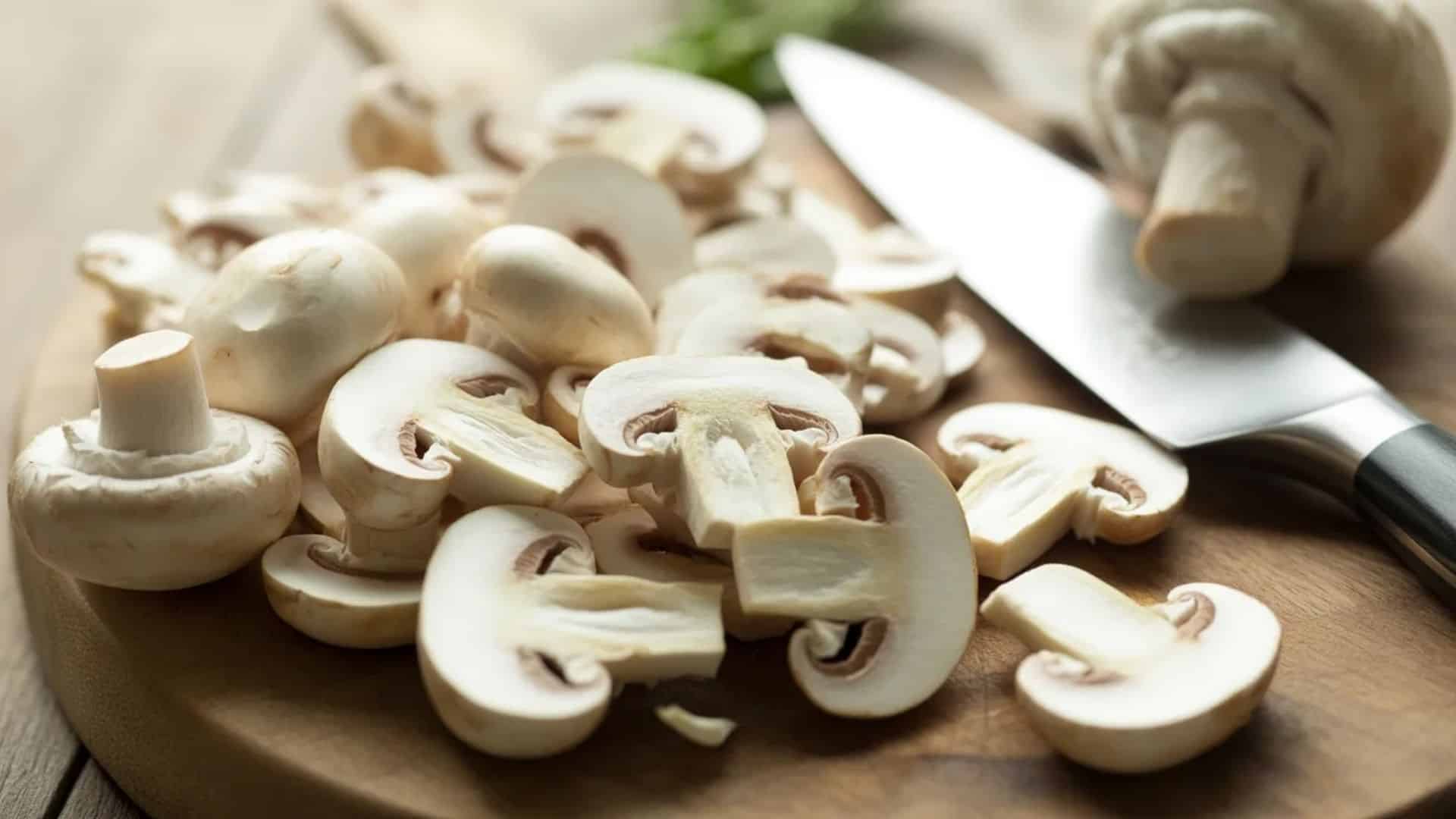 chopped white button mushrooms on a wooden cutting board with a kitchen knife beside them, close up food preparation scene
