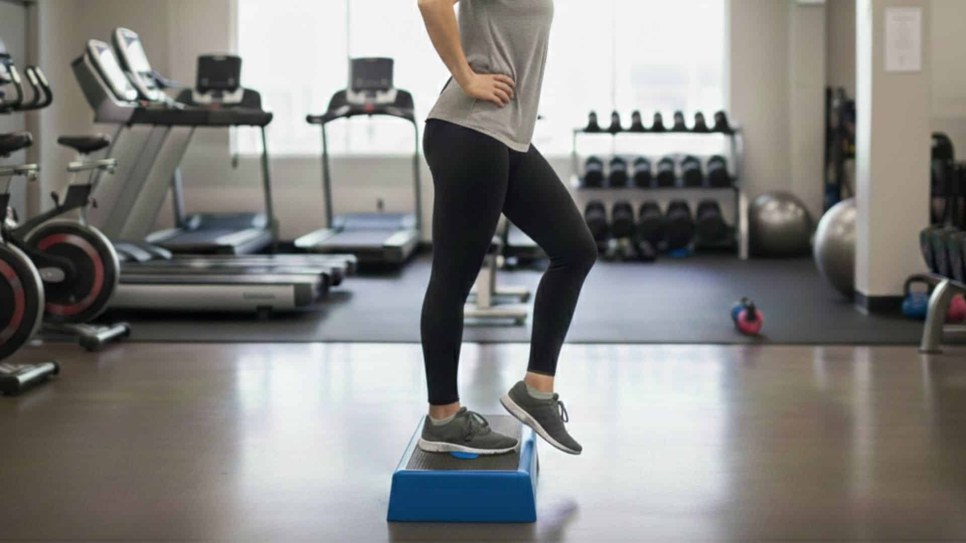 woman performing a hip drop exercise on a step platform in a gym, standing on one leg with the other lowered toward the floor